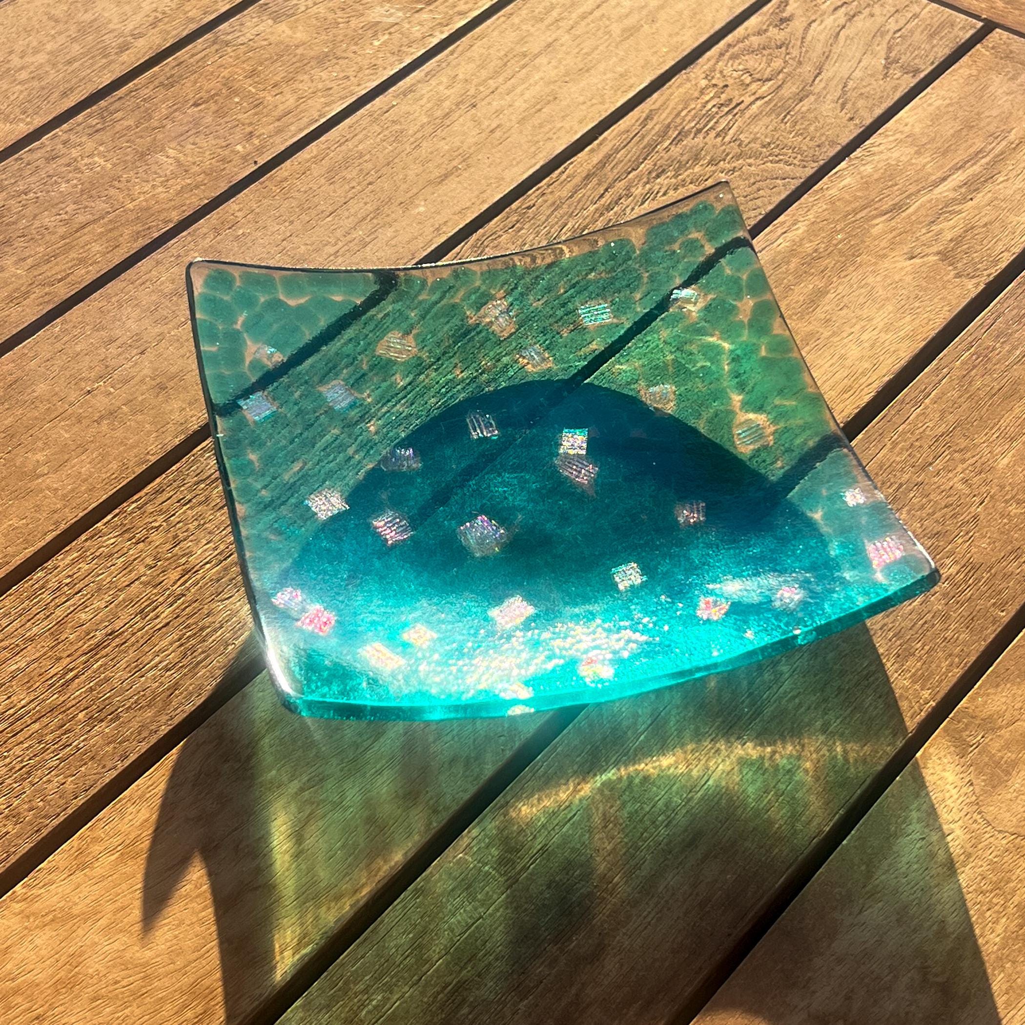 Six inch square dish made with bits transparent turquoise blue glass melted onto clear glass with random pieces of dichroic glass in the mix. Corners are slightly upturned. Dish is placed on a wood slat table in the sun with reflections.