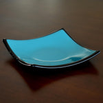 Handmade fused glass dish with a smooth glossy turquoise surface framed by sleek black edges. The shallow square shape curves upward at the corners, creating a modern yet simple decorative piece that reflects light beautifully against the dark wood background.