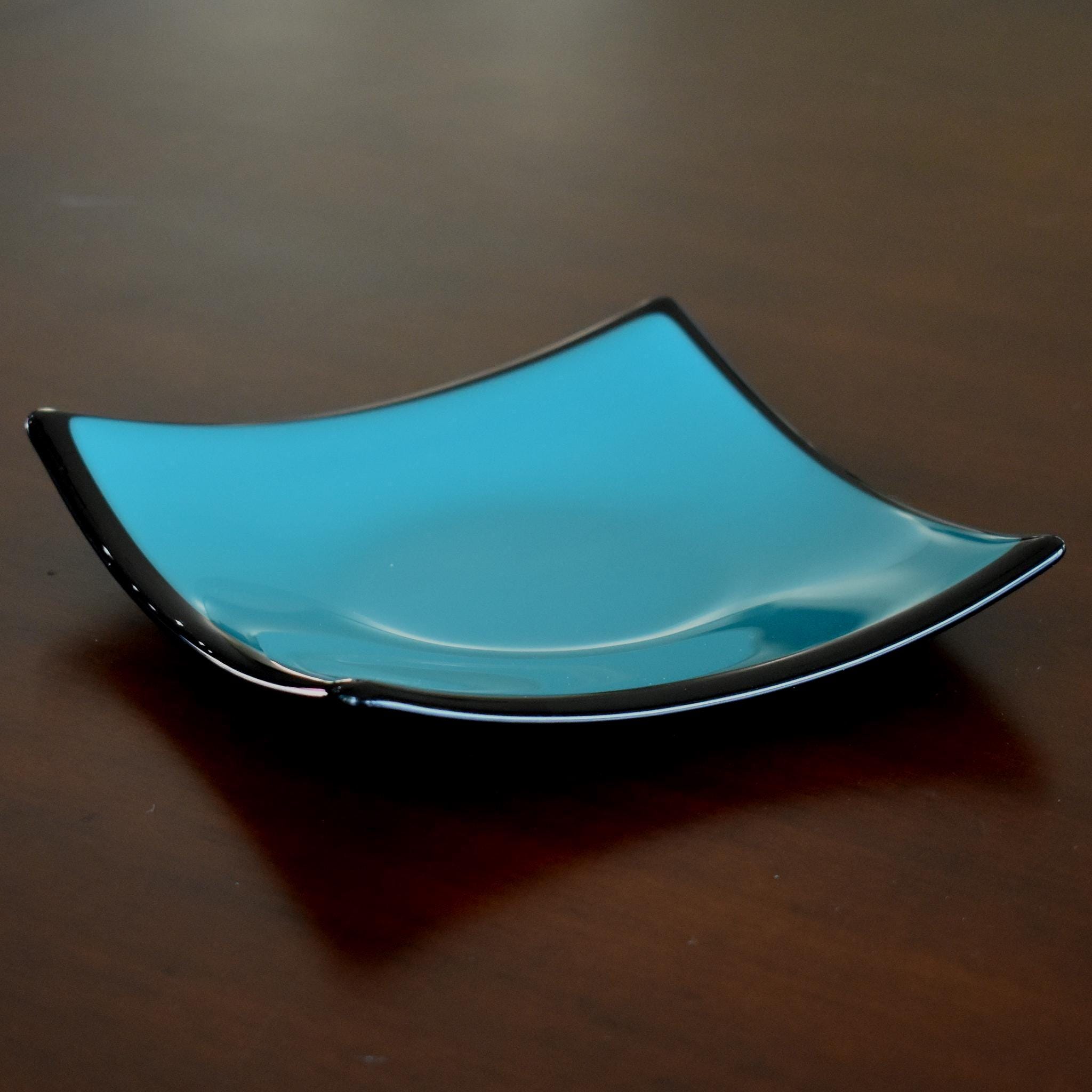 Handmade fused glass dish with a smooth glossy turquoise surface framed by sleek black edges. The shallow square shape curves upward at the corners, creating a modern yet simple decorative piece that reflects light beautifully against the dark wood background.
