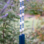 A hanging wind chime with two strings of white and blue beads, and two pieces of dark blue glass at the bottom. The beads are interspersed with small blue beads. The wind chime is suspended against a backdrop of green foliage.