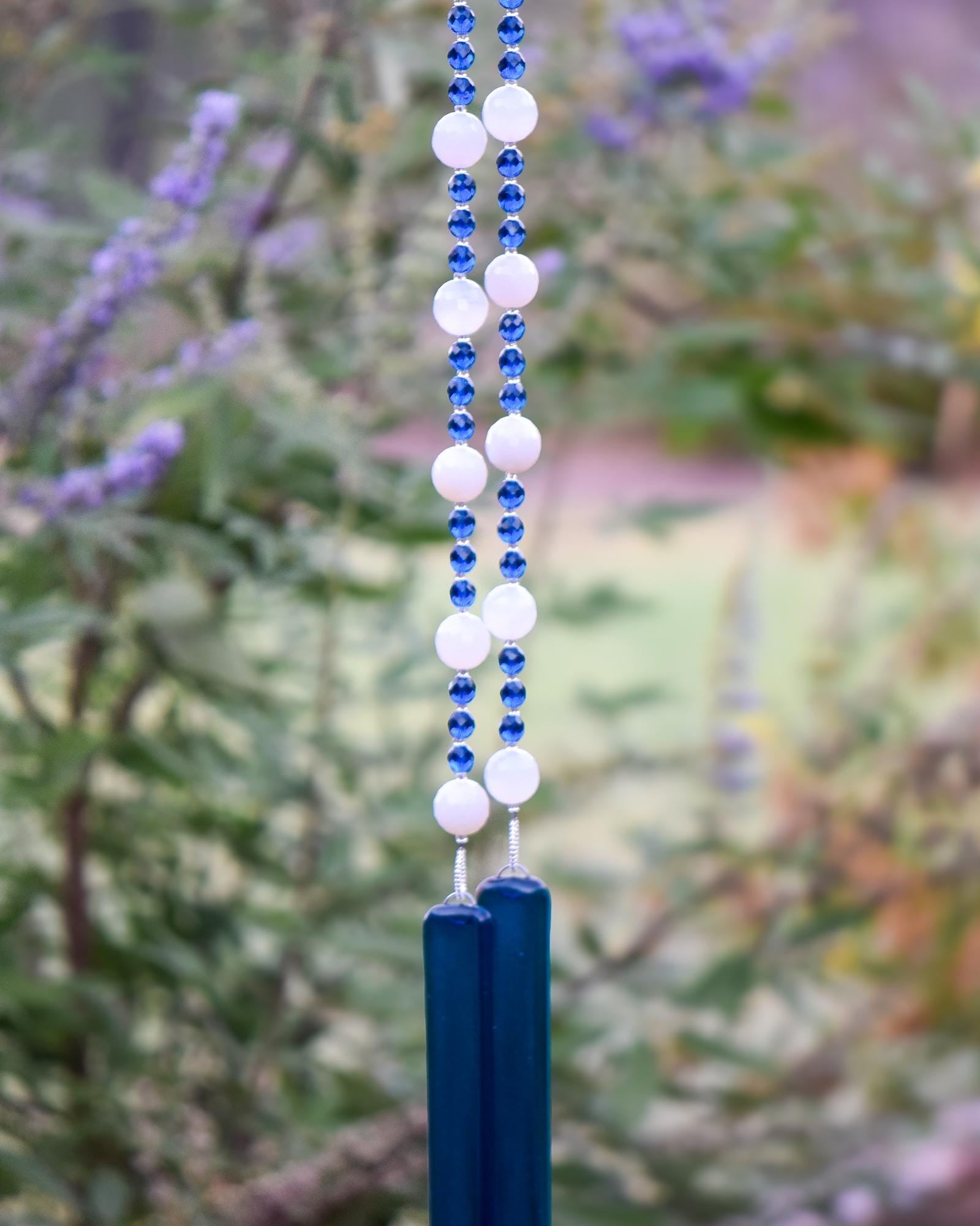 A hanging wind chime with two strings of white and blue beads, and two pieces of dark blue glass at the bottom. The beads are interspersed with small blue beads. The wind chime is suspended against a backdrop of green foliage.