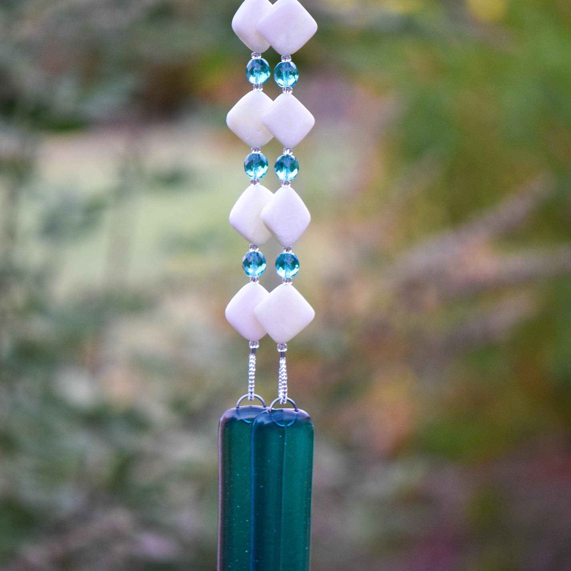 Handmade wind chime with white diamond-shaped beads, aqua crystal beads, and teal glass for garden or patio décor hanging vertically against foliage background.