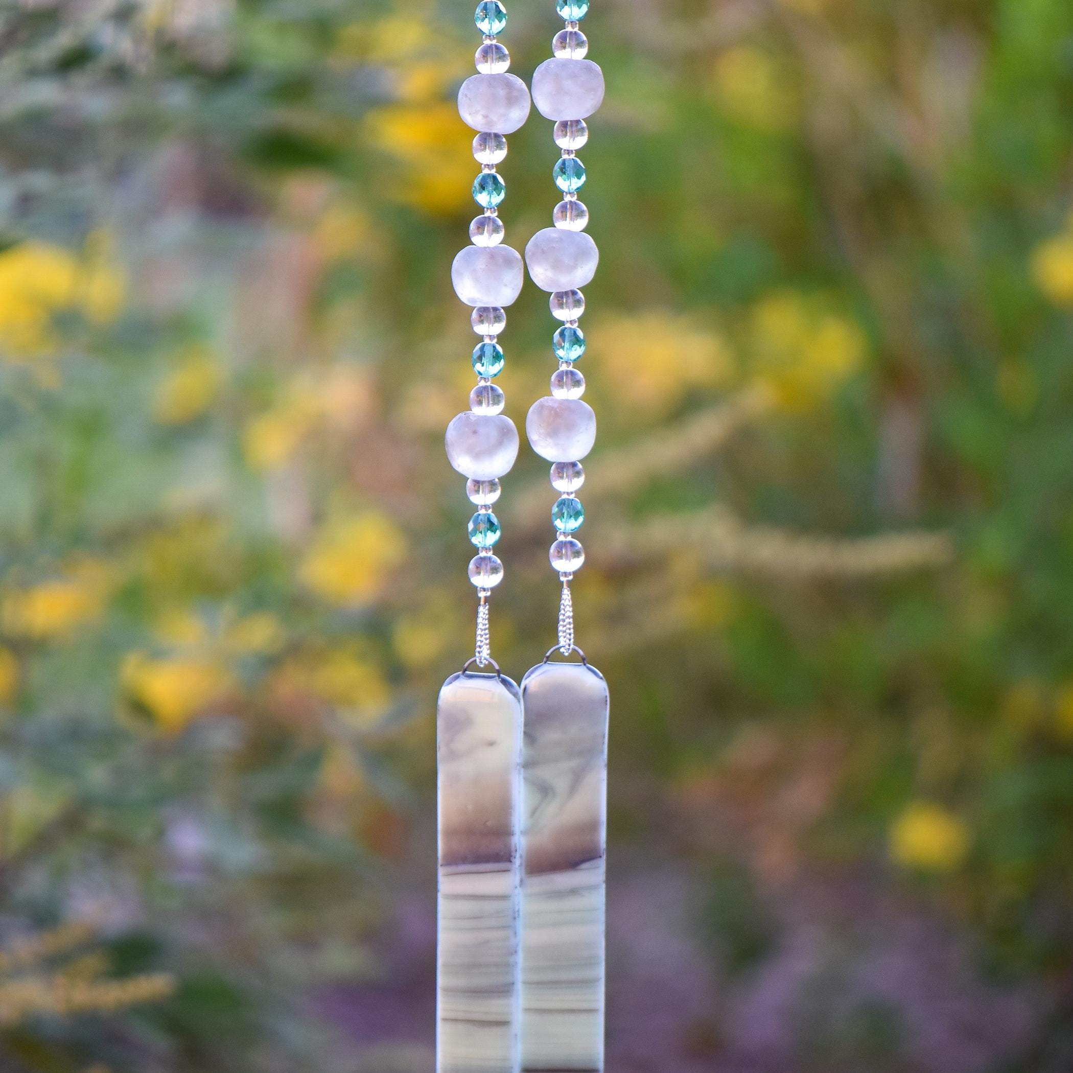 Handmade glass wind chime featuring smoky gray Krobo beads, aqua crystal accents, and two long fused glass pieces in soft neutral tones against blurred landscape background.