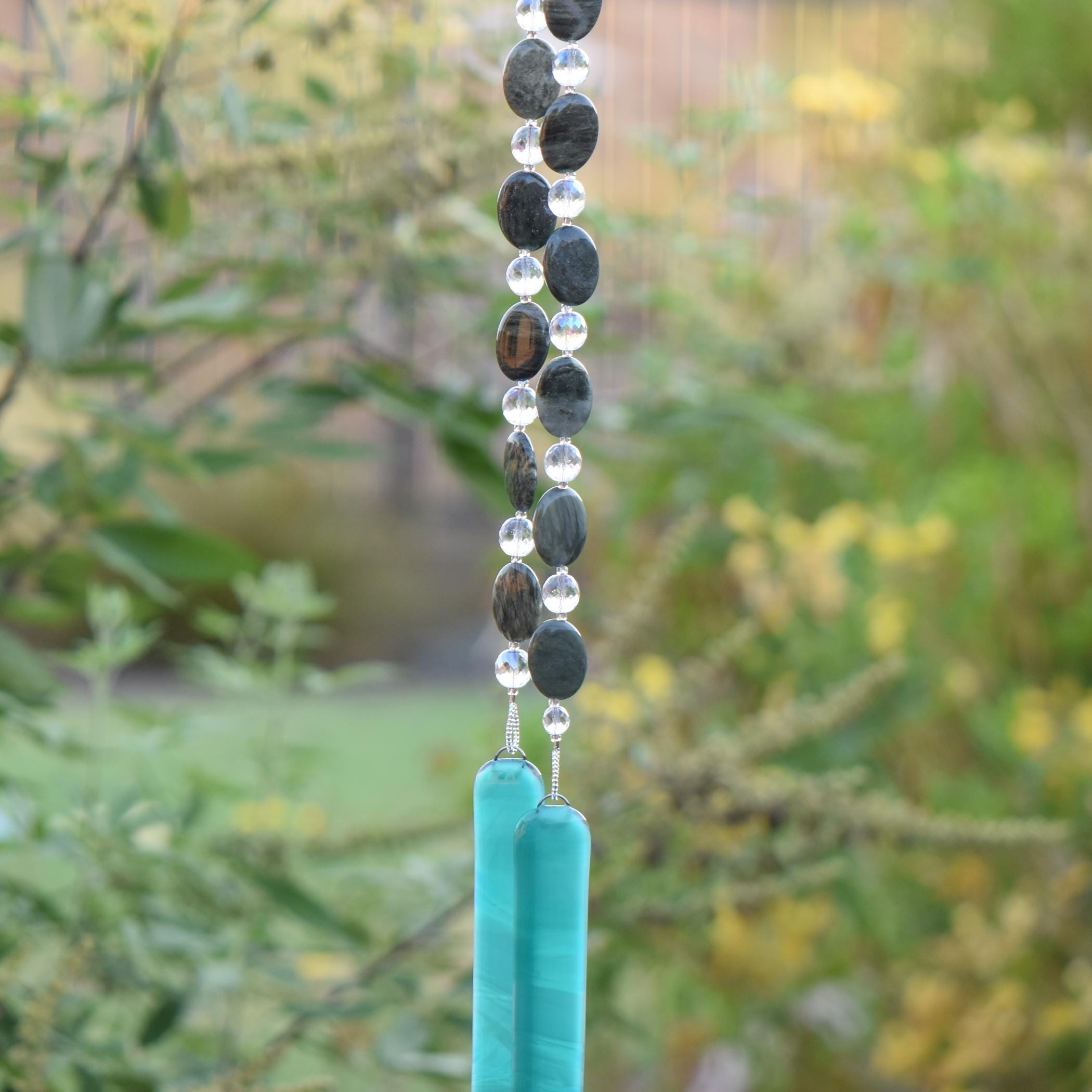 Large gray-green oval stone beads strung with clear glass lamp worked beads, hanging in two strands vertically; anchored by two pieces of teal and white fused glass, against blurred landscape background.