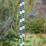 A hanging wind chime with two strings of white and blue beads. The beads are interspersed with small blue beads. The wind chime is suspended against a backdrop of green foliage.