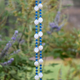 A hanging wind chime with two strings of white and blue beads. The beads are interspersed with small blue beads. The wind chime is suspended against a backdrop of green foliage.
