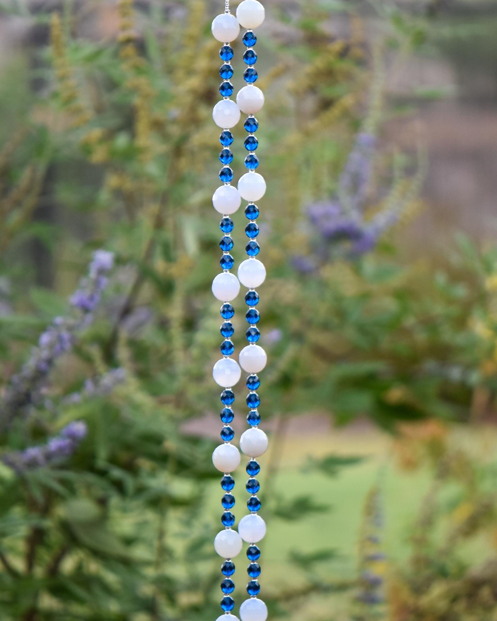 A hanging wind chime with two strings of white and blue beads. The beads are interspersed with small blue beads. The wind chime is suspended against a backdrop of green foliage.