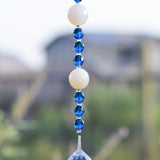 Long sun catcher from Cinjo Handmade hanging against blurred background. Two large white quartz beads are shown with 3 dark blue beads in between each of the quartz beads. A round glass crystal prism anchors the piece.