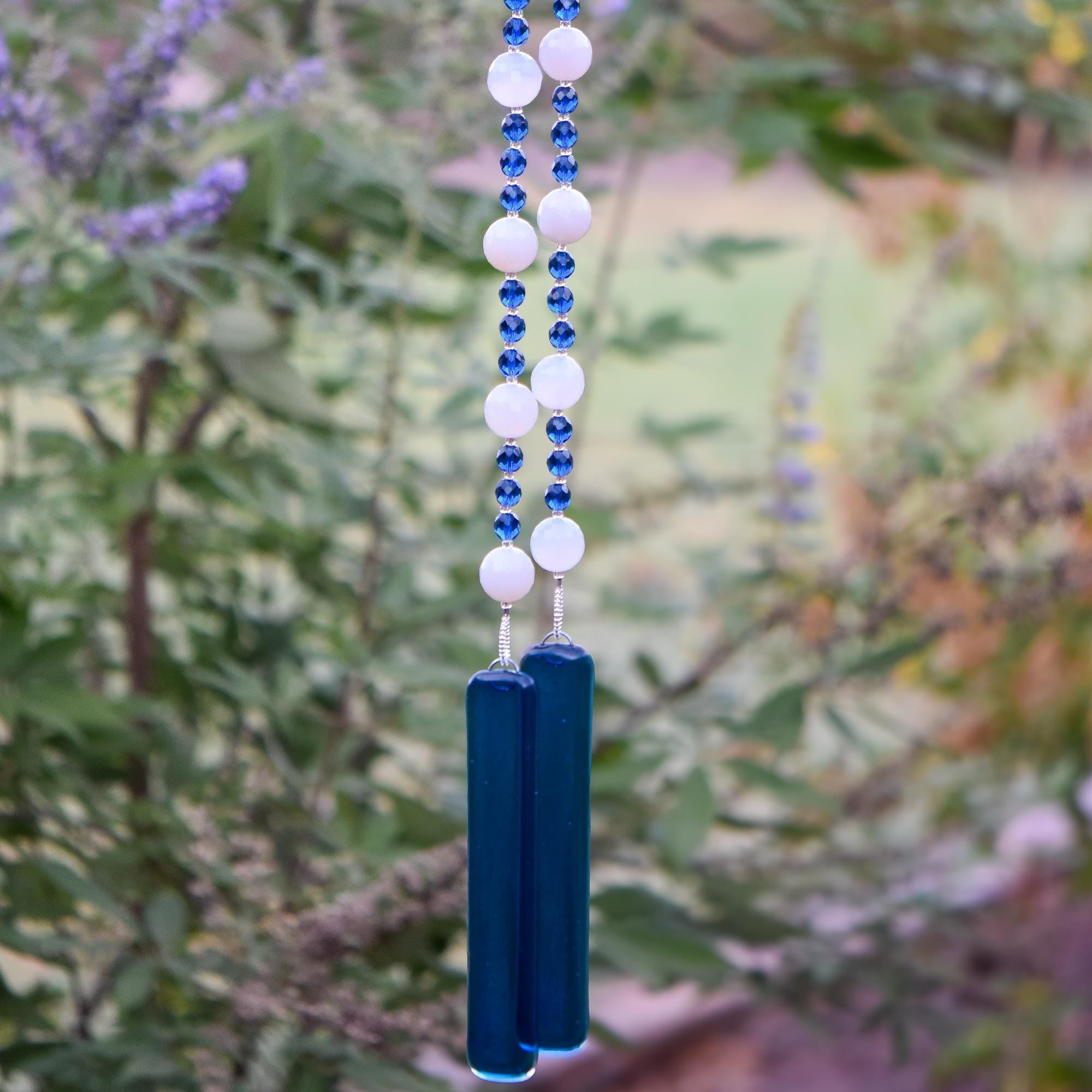 Large, round white quartz stone beads strung with dark aqua blue glass beads, hanging vertically in two strands, anchored by two pieces of dark blue fused glass, against foliage background.