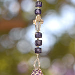 Small 9&quot; beaded sun catcher hanging vertically against blurred foliage background. Sun catcher is made with six Murano glass beads in purple with silver foil flanking a faceted glass crystal cross, anchored by a very large sterling silver pendant made with swirls of purple crystals.