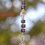 Small 9&quot; beaded sun catcher hanging vertically against blurred foliage background. Sun catcher is made with six Murano glass beads in purple with silver foil flanking a faceted glass crystal cross, anchored by a very large sterling silver pendant made with swirls of purple crystals.