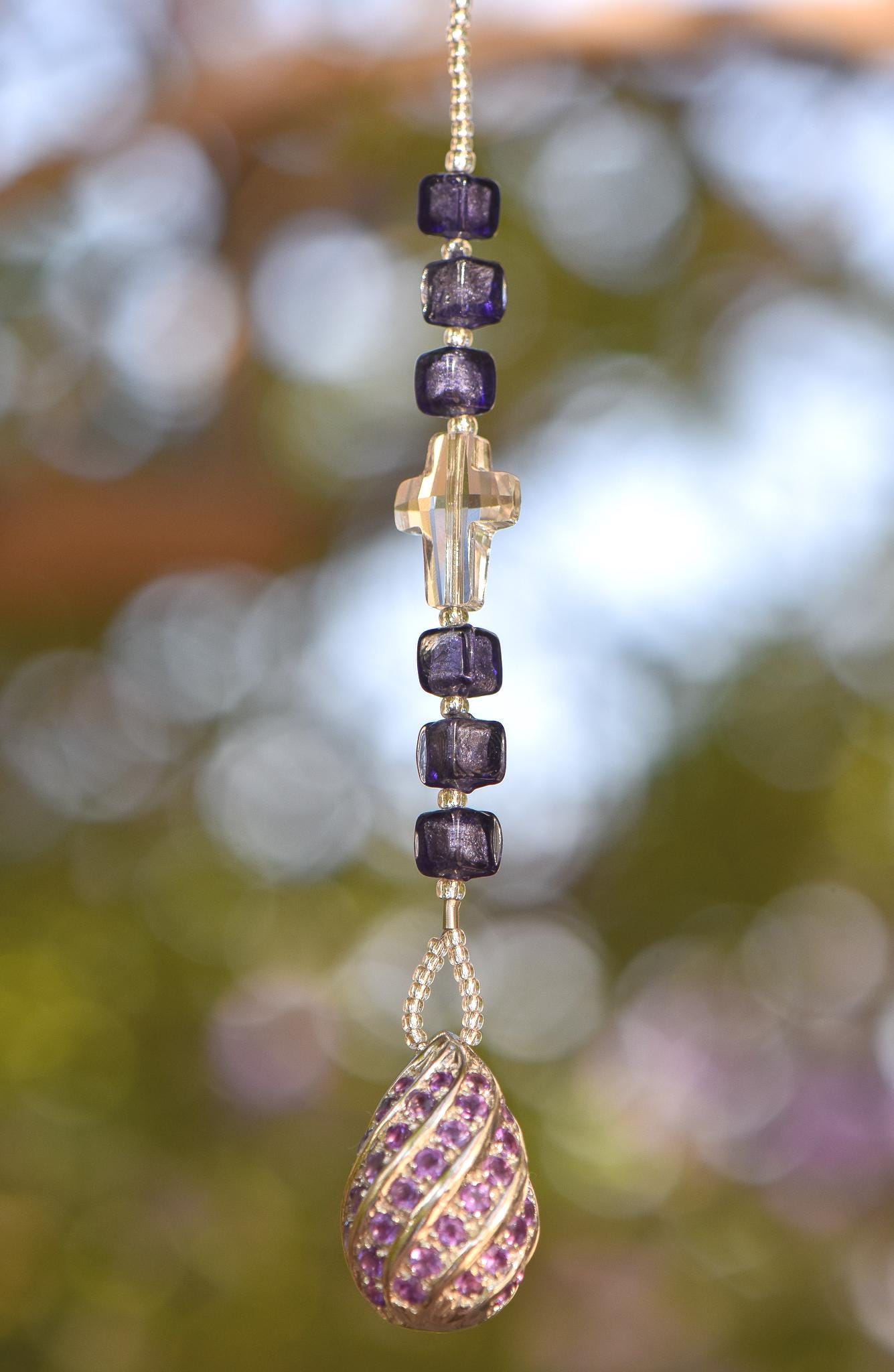 Small 9&quot; beaded sun catcher hanging vertically against blurred foliage background. Sun catcher is made with six Murano glass beads in purple with silver foil flanking a faceted glass crystal cross, anchored by a very large sterling silver pendant made with swirls of purple crystals.