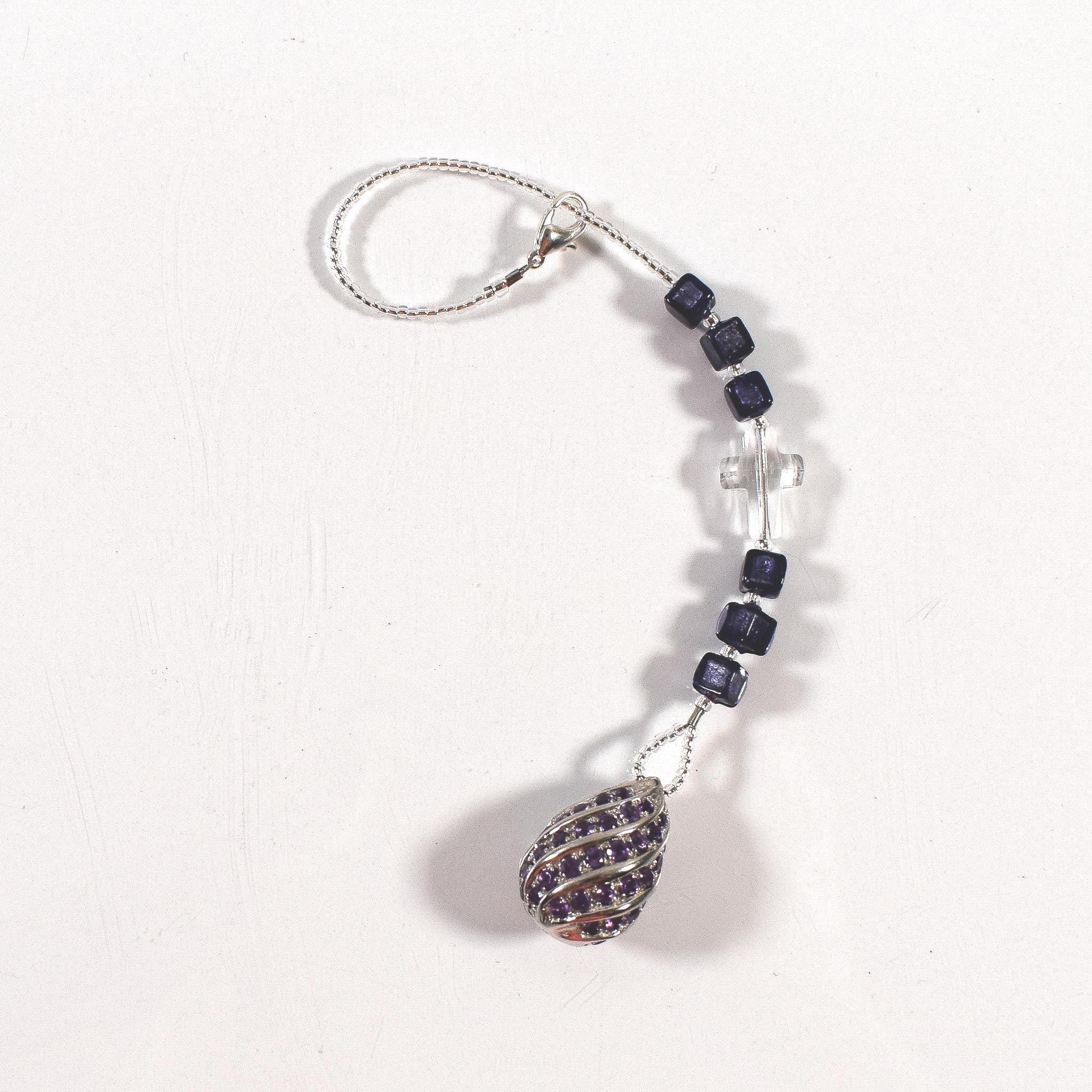 Small purple murano glass sun catcher laying flat on white background. Sun catcher has six purple murano cube beads flanking a glass crystal cross and anchored by a large sterling silver pendant. At the top of the sun catcher is a seed bead-covered wire ending in a silver plated clasp