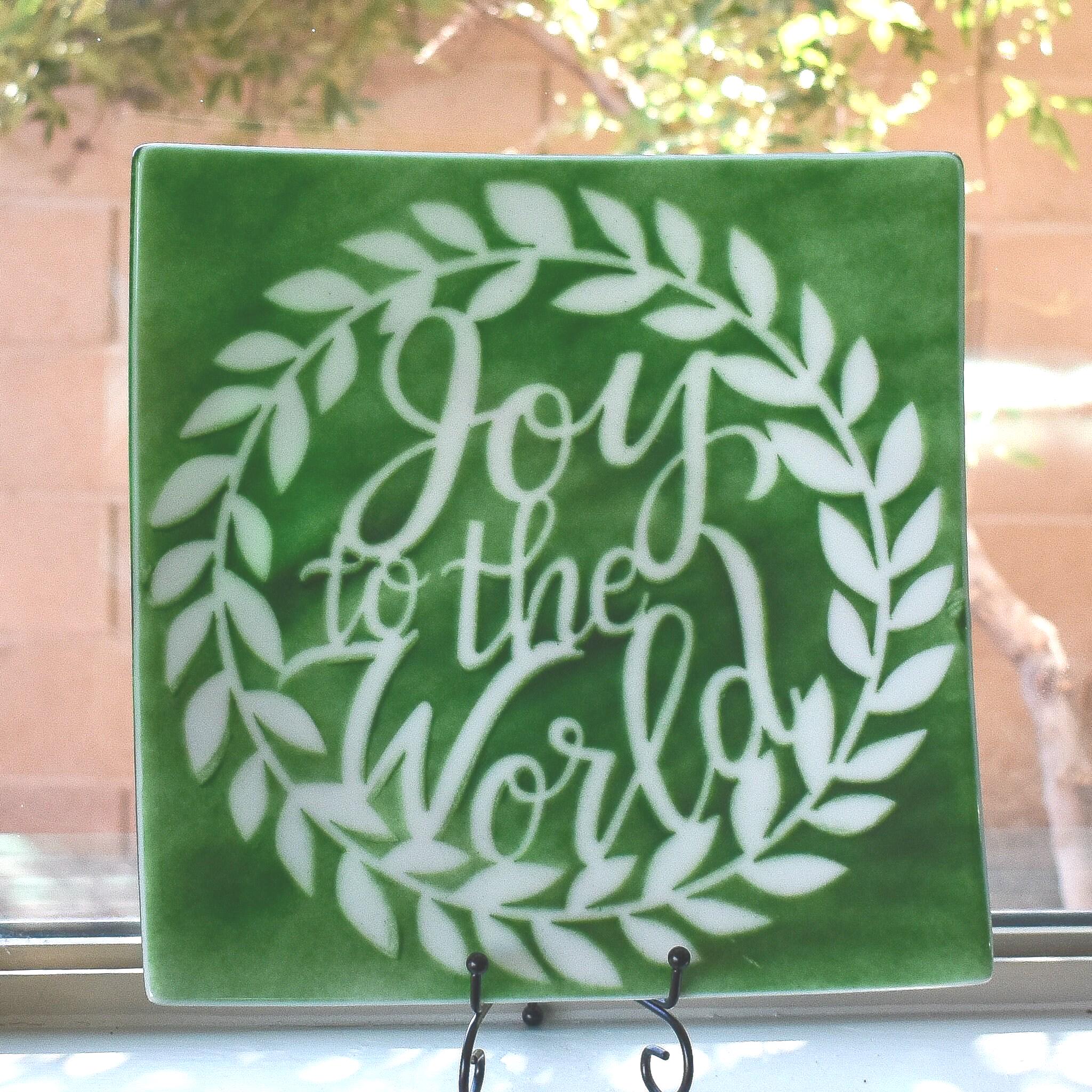 Large square fused glass platter in green with white lettering on stand in window.