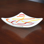 Six inch square dish in creamy white with abstract design of orange, red and yellow fused glass on dark brown wood surface, from the side.