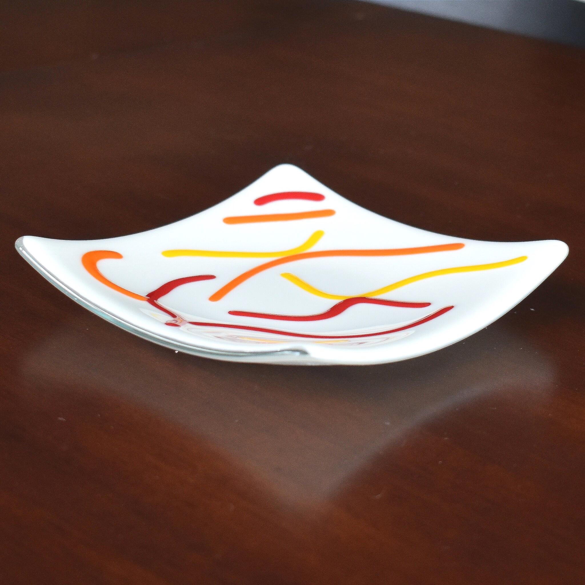 Six inch square dish in creamy white with abstract design of orange, red and yellow fused glass on dark brown wood surface, from the side.