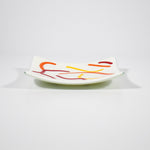 Small, square fused glass tray is cream with orange, red and yellow curve design against white background, view slightly from the side.