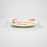 Small, square fused glass tray is cream with orange, red and yellow curve design against white background, view slightly from the side.