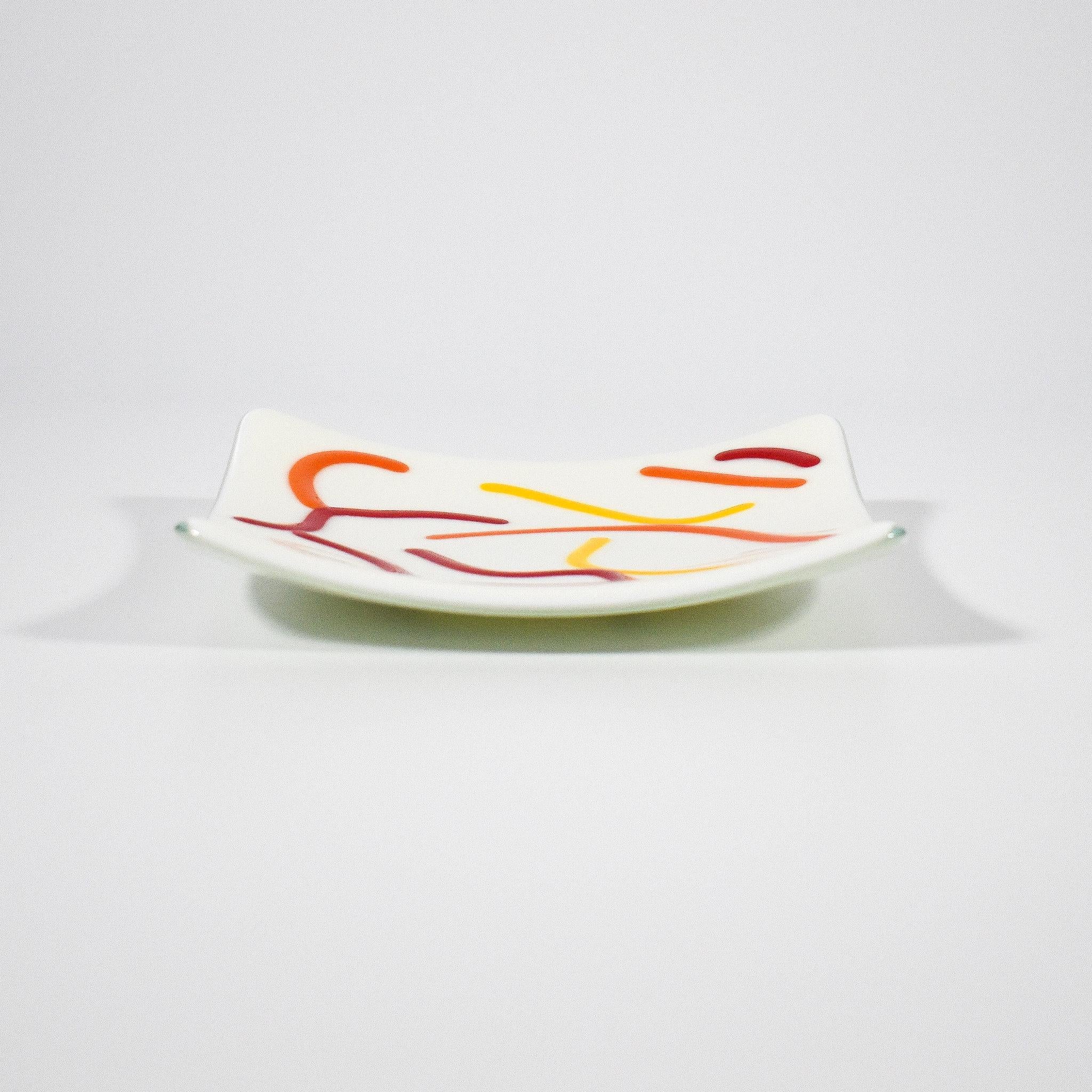 Small, square fused glass tray is cream with orange, red and yellow curve design against white background, view slightly from the side.