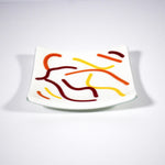 Square glass tray with vanilla cream basis and abstract design of red, orange and yellow, against white background.