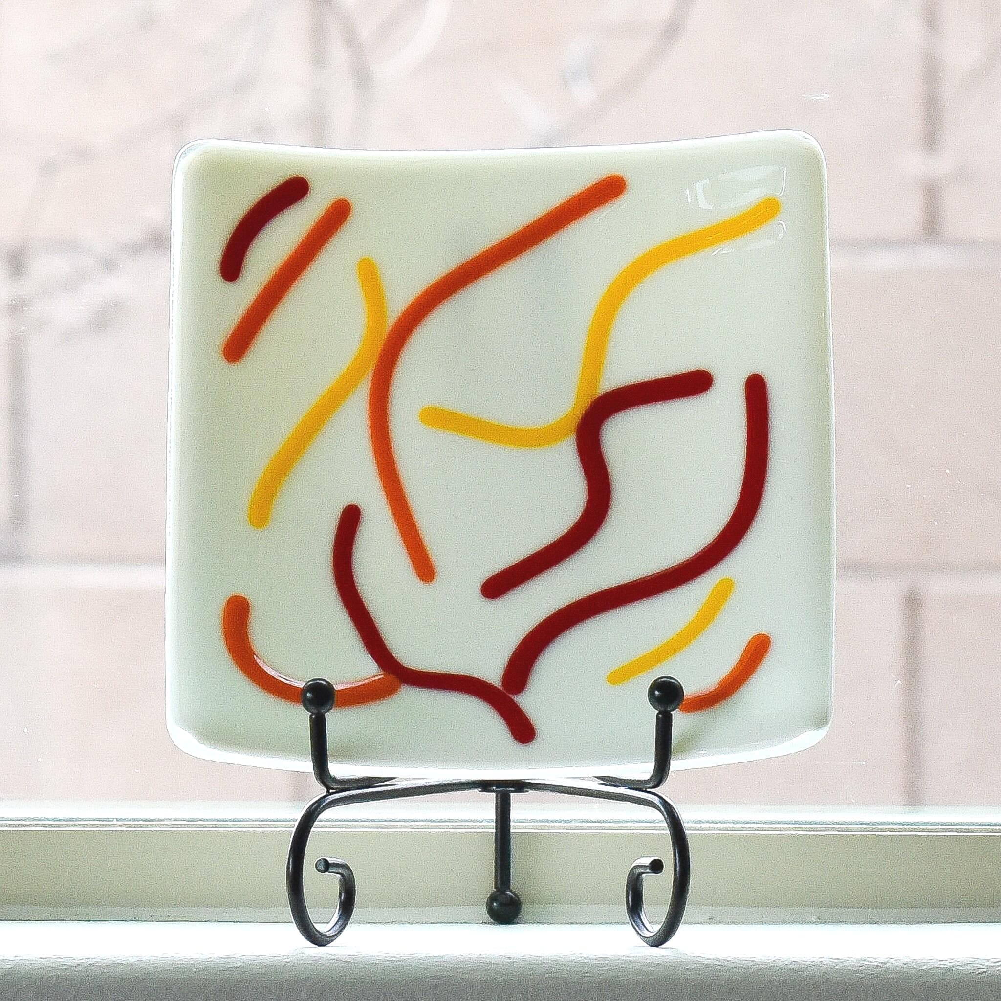 Square glass tray with vanilla cream basis and abstract design of red, orange and yellow, on stand, in window.