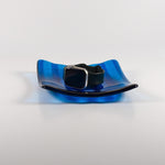 Deep aqua blue fused glass tray with two opposite scalloped edges and strips of a lighter color. The photo is looking at the front of the tray and the tray is on a white surface. The tray is holding a watch to show size and use.