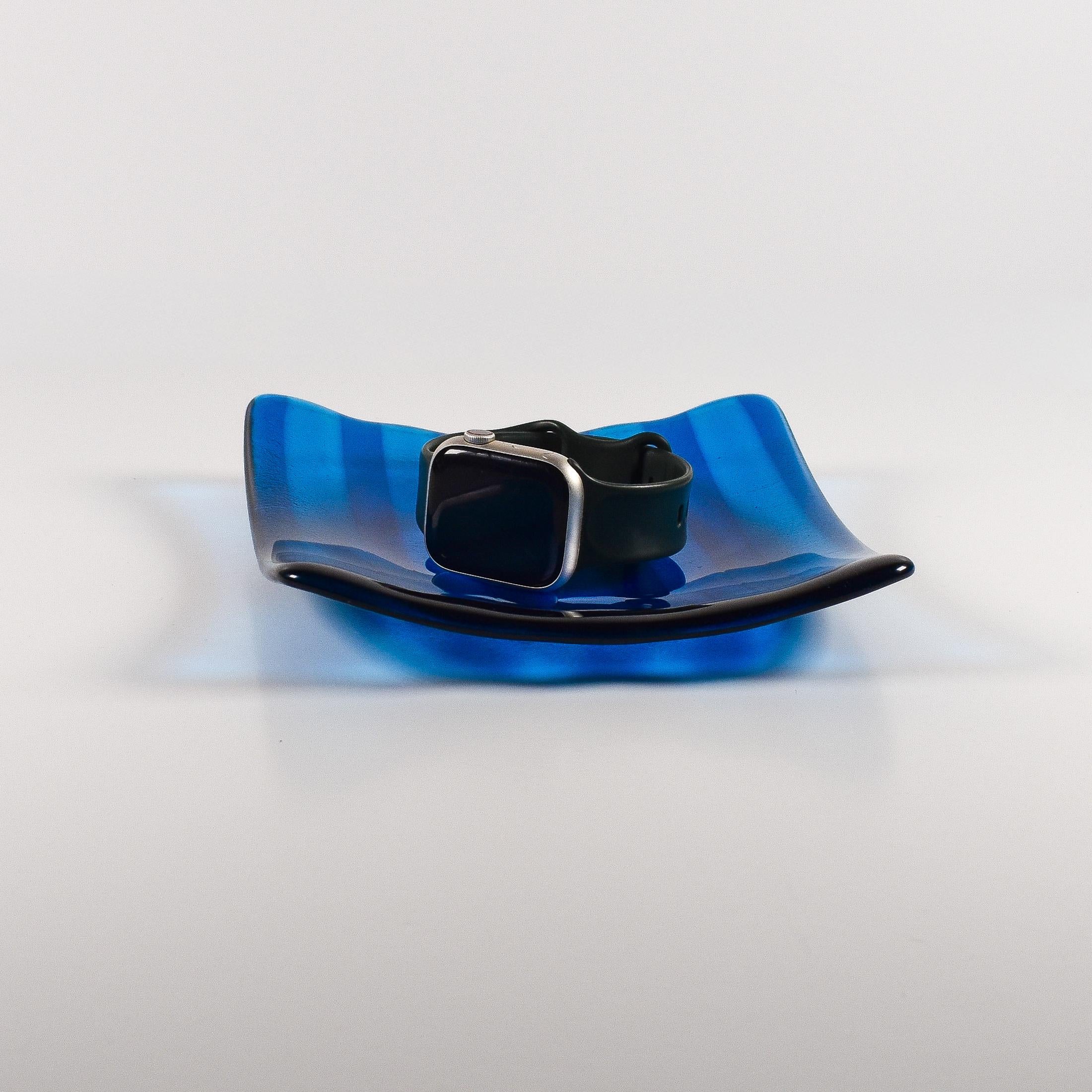 Deep aqua blue fused glass tray with two opposite scalloped edges and strips of a lighter color. The photo is looking at the front of the tray and the tray is on a white surface. The tray is holding a watch to show size and use.