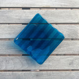 Deep aqua blue fused glass tray with two opposite scalloped edges and strips of a lighter color. The photo is looking down from the top of the tray. The tray is sitting on a wood table, outdoors, in shade, with some reflections in the glass.