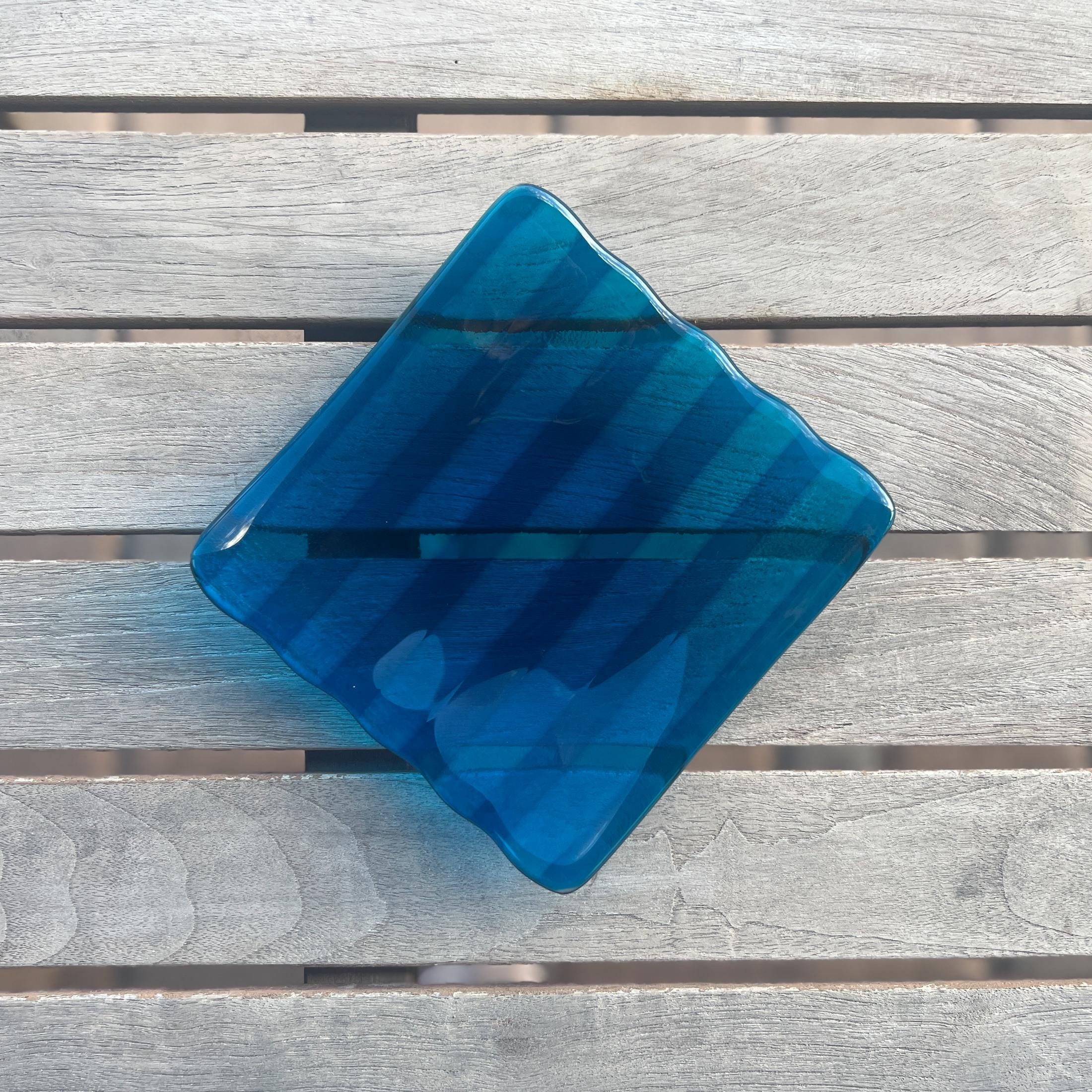 Deep aqua blue fused glass tray with two opposite scalloped edges and strips of a lighter color. The photo is looking down from the top of the tray. The tray is sitting on a wood table, outdoors, in shade, with some reflections in the glass.
