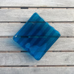 Deep aqua blue fused glass tray with two opposite scalloped edges and strips of a lighter color. The photo is looking down from the top of the tray. The tray is sitting on a wood table, outdoors, in shade, with some reflections in the glass.