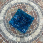 Deep aqua blue fused glass tray with two opposite scalloped edges and strips of a lighter color. The photo is looking down from the top of the tray. The tray is sitting on a stone table that has a geometric design that shows through the glass. Photo was taken outdoors and there is some sunlight reflection on the glass.