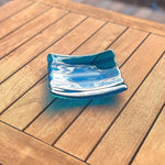 Deep aqua blue fused glass tray with two scalloped edges and strips of a lighter color. The tray is sitting on a wood table at an angle, outdoors, in sunlight, with some reflections in the glass.