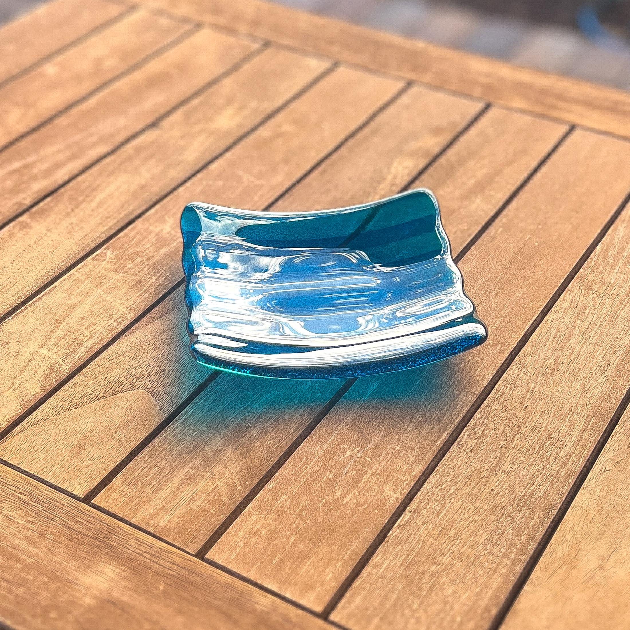 Deep aqua blue fused glass tray with two scalloped edges and strips of a lighter color. The tray is sitting on a wood table at an angle, outdoors, in sunlight, with some reflections in the glass.