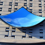 Square fused glass dish with solid blue center, black trim and black base, sitting on iron mesh on stone table.