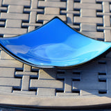 Square fused glass dish with solid blue center, black trim and black base, sitting on iron mesh on stone table.