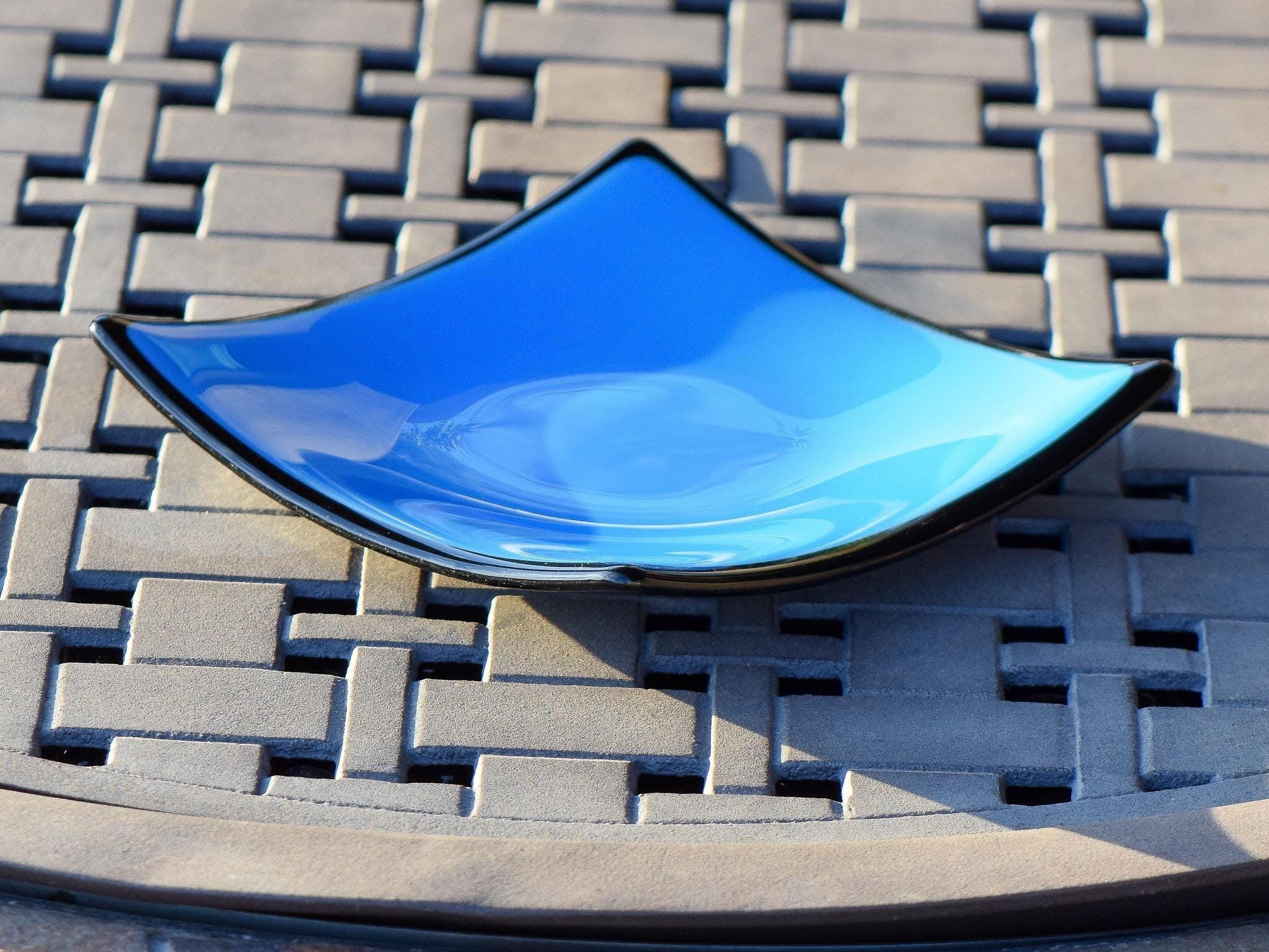 Square fused glass dish with solid blue center, black trim and black base, sitting on iron mesh on stone table.