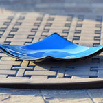 Side view of square fused glass dish with solid blue center, black trim and black base, sitting on iron mesh on stone table.