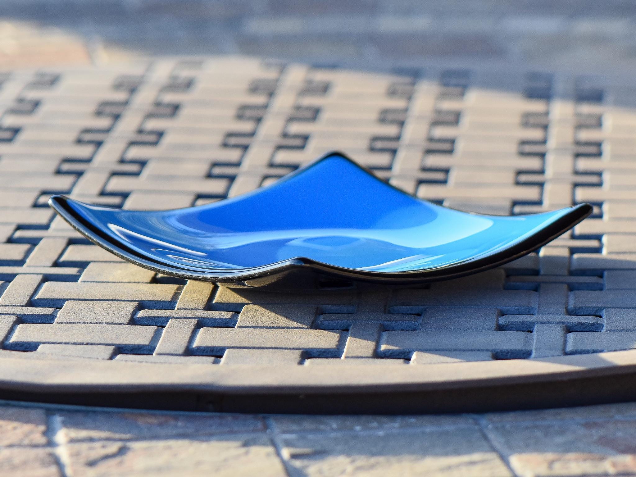Side view of square fused glass dish with solid blue center, black trim and black base, sitting on iron mesh on stone table.