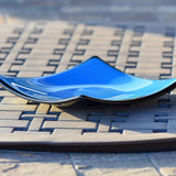 Side view of square fused glass dish with solid blue center, black trim and black base, sitting on iron mesh on stone table.