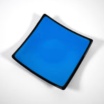 Square fused glass dish with solid blue center, black trim and black base, sitting white surface.