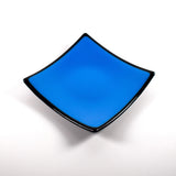 Square fused glass dish with solid blue center, black trim and black base, sitting white surface.