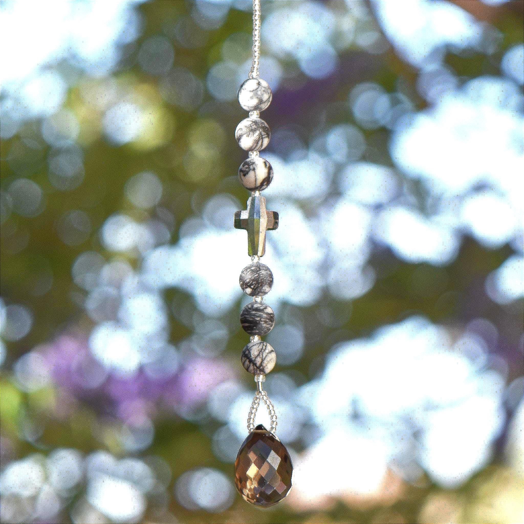 Small sun catcher hanging vertically against blurred foliage background. Silver-gray crystal cross is flanked by six jasper stone beads in a marbled gray, anchored by large smoky quartz pendant