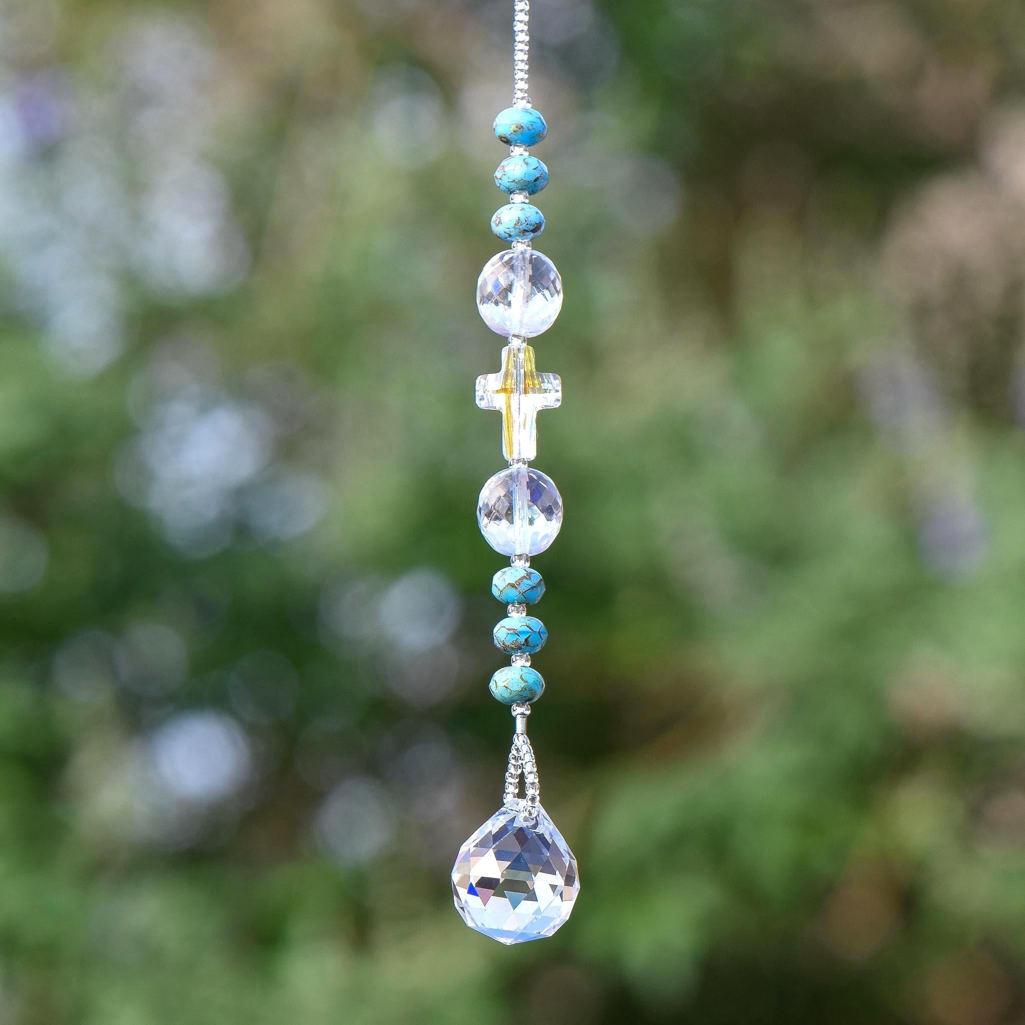 9 inch long sun catcher hanging vertically against blurred foliage background, consisting of a single glass crystal cross bed with reflective finish, flanked by two large round glass crystal beads and six small rondelle-shaped turquoise beads, all anchored by a small glass crystal prism.