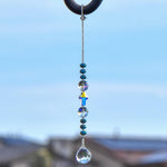 9 inch long sun catcher hanging vertically against sky background, consisting of a single glass crystal cross bed with reflective finish, flanked by two large round glass crystal beads and six small rondelle-shaped turquoise beads, all anchored by a small glass crystal prism.