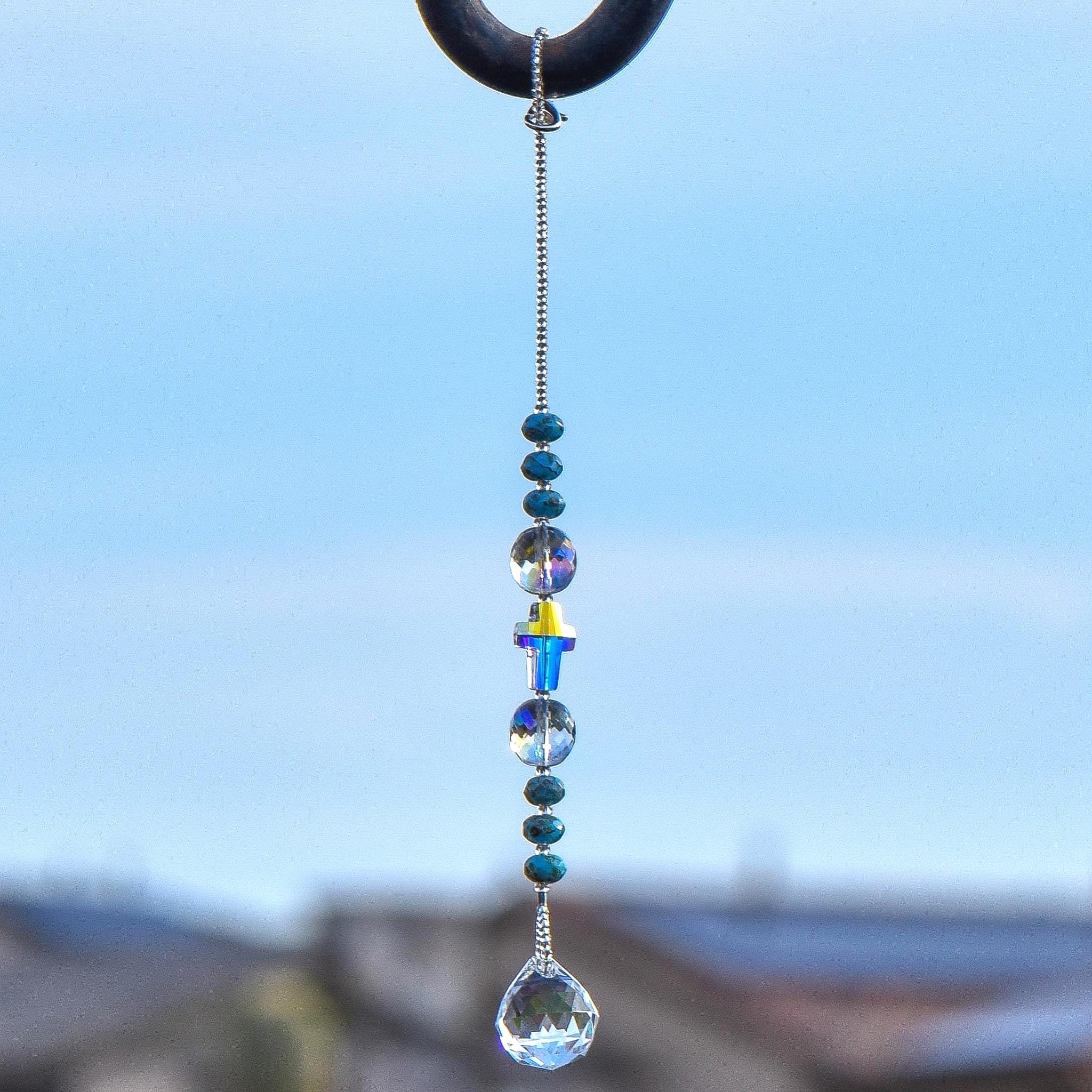 9 inch long sun catcher hanging vertically against sky background, consisting of a single glass crystal cross bed with reflective finish, flanked by two large round glass crystal beads and six small rondelle-shaped turquoise beads, all anchored by a small glass crystal prism.