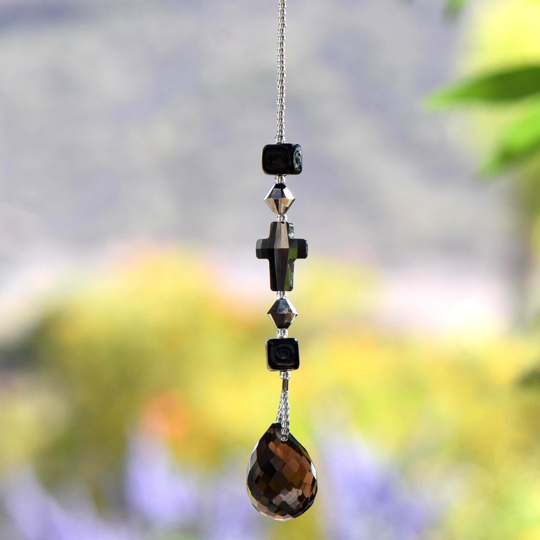 Gray Silver glass crystal cross beaded with two gray silver bicone beads and 2 Murano cubic black glass beads, hanging vertically against blurred background. A smoky quartz pendant serves as an anchor for the sun catcher.