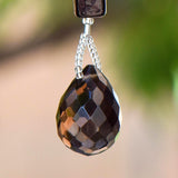 A teardrop-shaped faceted, dark brown smoky quartz gemstone. The gem hangs from a silver-colored chain of small beads, connected to a rectangular, black bead. The background is blurred, with hints of green and beige.
