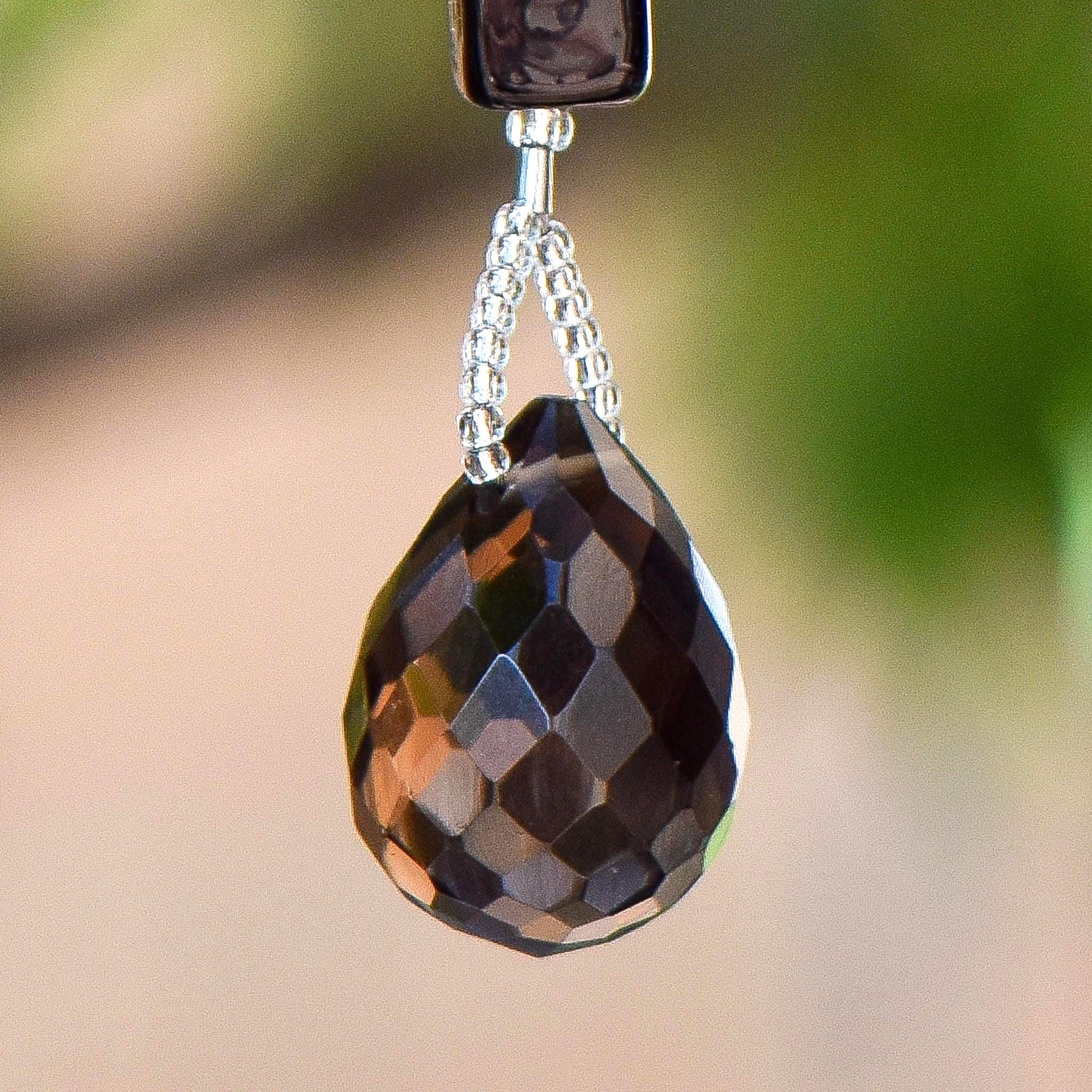 A teardrop-shaped faceted, dark brown smoky quartz gemstone. The gem hangs from a silver-colored chain of small beads, connected to a rectangular, black bead. The background is blurred, with hints of green and beige.