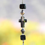 Gray Silver glass crystal cross beaded with two gray silver bicone beads and 2 Murano cubic black glass beads, hanging vertically against blurred background.