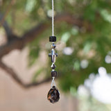 Gray Silver glass crystal cross beaded with two gray silver bicone beads and 2 Murano cubic black glass beads, hanging vertically against blurred background.